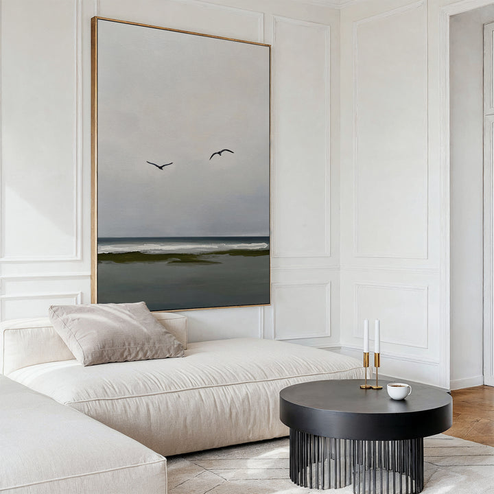 A modern, minimalist living room features a cream sofa, black round coffee table, candles, cup, and serene decor highlighted by the Freeflight Canvas Art—large wall art depicting an ocean view with two birds flying.