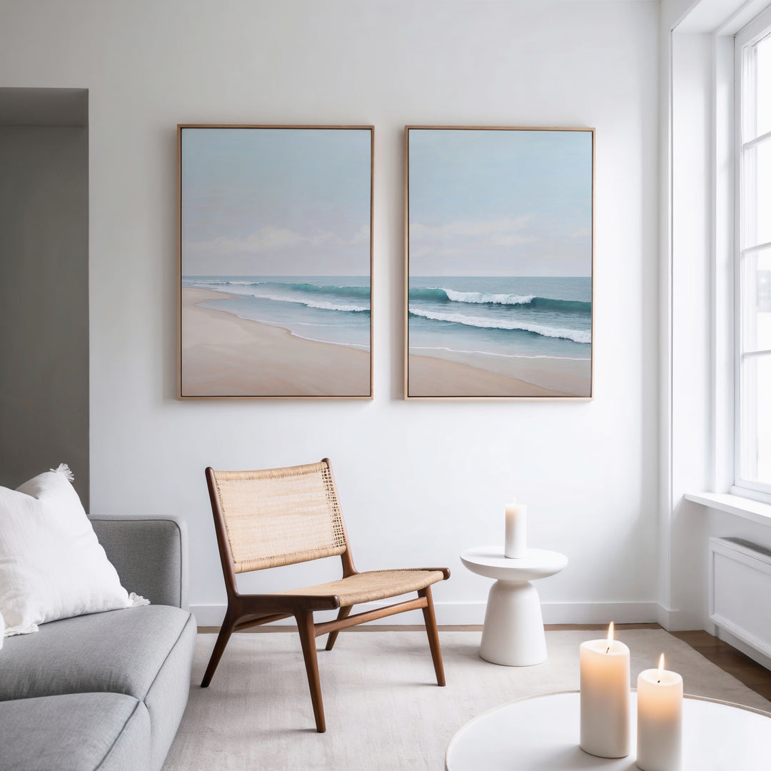 A bright, modern living room features a light gray sofa, woven chair, small white side table, and three lit candles. Above the chair hang two framed Gentle Surf Set of 2 coastal art pieces, adding charm as sunlight streams through tall windows.