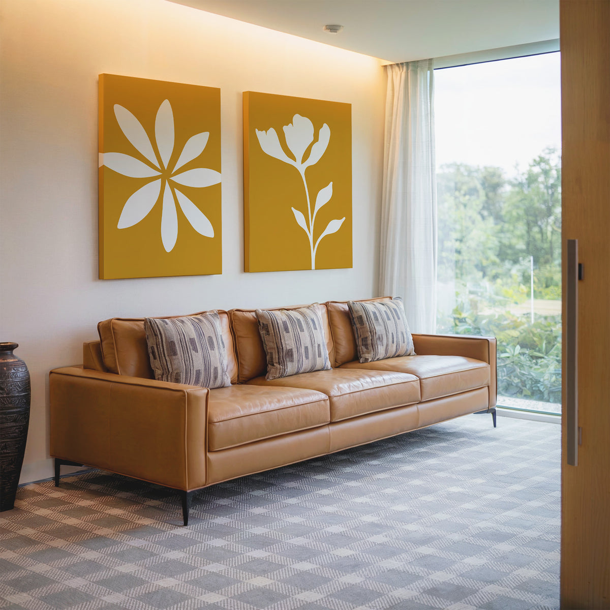 A modern living room featuring a tan leather sofa with four patterned pillows, the Golden Bloom Set of 2 yellow floral wall art above, a dark vase nearby, and a large window with sheer curtains overlooking greenery.