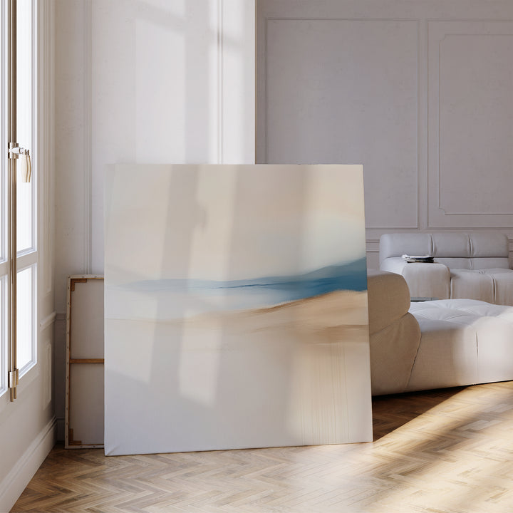The Golden Drift Canvas Art, featuring soft beige and blue tones, rests against a white wall beside a modern white sofa and tall windows—ideal for adding contemporary style to your sunlit living room with parquet flooring.