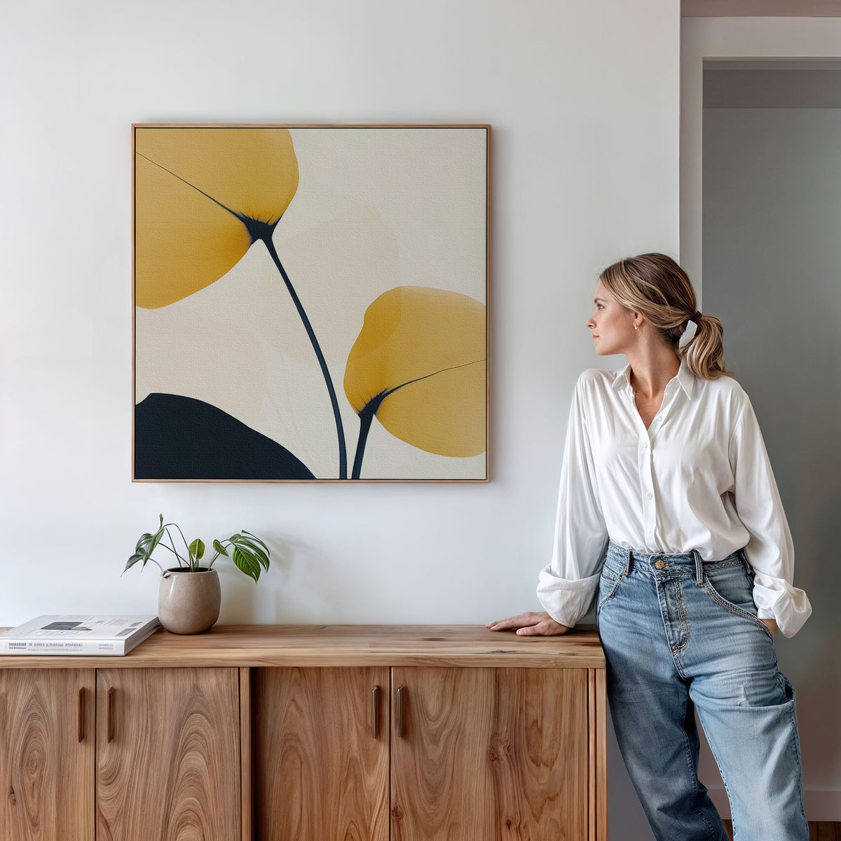 A woman in a white blouse and jeans leans against a wooden cabinet, admiring the Golden Leaves Canvas Art—minimalist wall art with yellow and black shapes—on a white wall. A small plant and books complete the stylish living room decor.