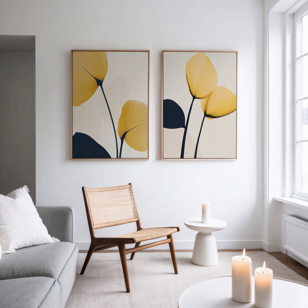 A modern living room features a gray sofa, a wooden chair with a woven seat, a small white side table, lit candles, and the Golden Leaves Set of 2 wall art in yellow and dark blue. Sunlight through a large window adds warmth to the decor.
