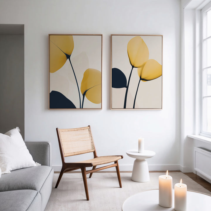 A modern living room features a gray sofa, a wooden chair with a woven seat, a small white side table, lit candles, and the Golden Leaves Set of 2 wall art in yellow and dark blue. Sunlight through a large window adds warmth to the decor.