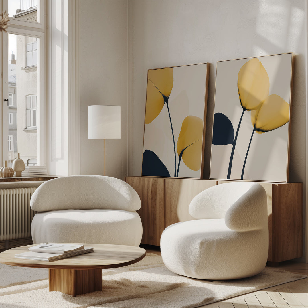 A modern living room features two white curved armchairs, a round wooden coffee table, and minimalist decor. The Golden Leaves Set of 2 wall art with yellow and navy leaves sits on a wooden sideboard in natural light.