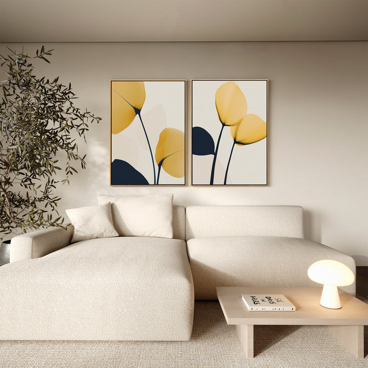 A modern living room with a cream sectional, a small wooden coffee table, a lamp, books, a large potted plant, and minimalist wall art—Golden Leaves Set of 2—featuring abstract yellow and navy botanicals completes the decor.