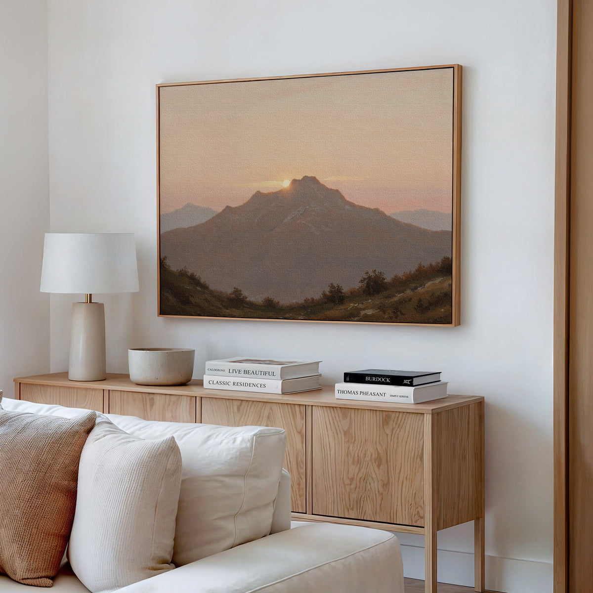 A cozy, modern living room features a light wood console, white lamp, decor accents, books, and the Golden Summit Canvas Art—a striking sunrise over mountains—hanging above to elevate your living room decor.