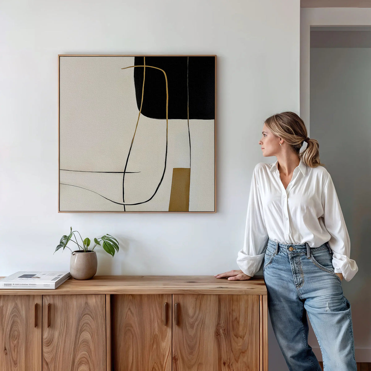 A woman in a white shirt and jeans leans against a wooden cabinet, admiring the Golden Thread Canvas Art—a modern abstract piece with black, white, and gold lines—while a small plant and books accentuate the minimalist decor.