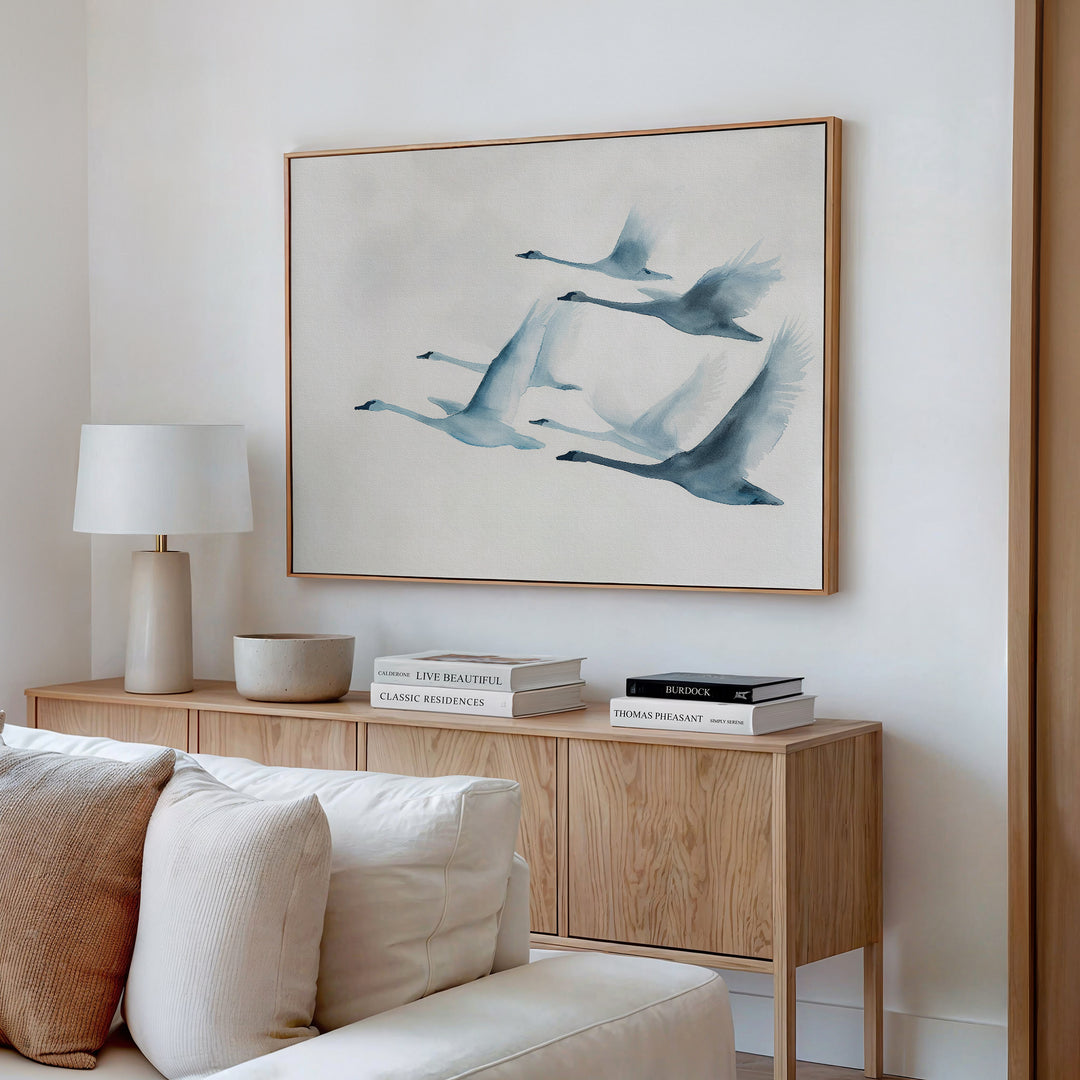 A minimalist living room features a wooden sideboard with books, a lamp, and decor beneath the Graceful Flock Canvas Art—a framed painting of five blue-toned birds. A cream sofa appears in the foreground, complementing this modern nature decor.