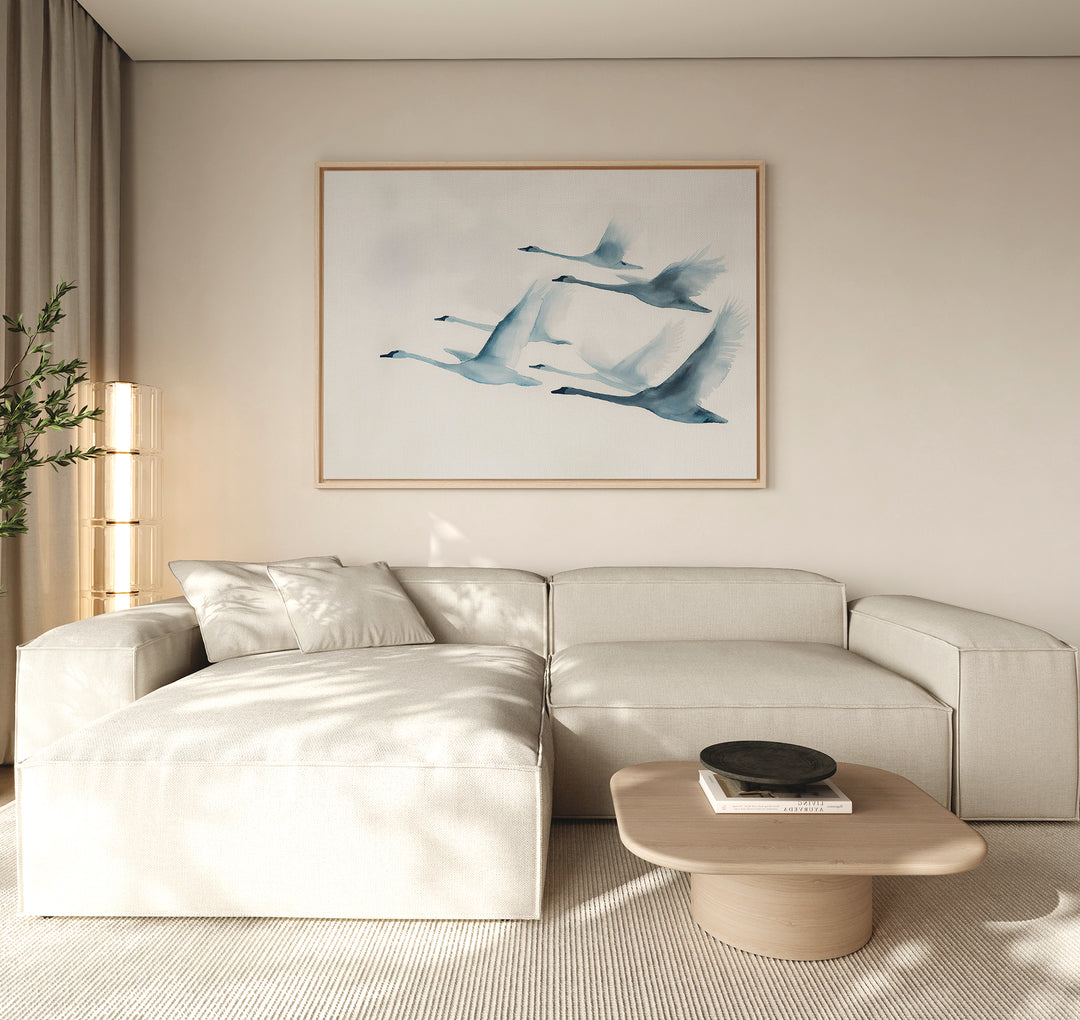 A modern living room features a light beige sectional sofa, a low wooden coffee table, and the Graceful Flock Canvas Art with flying blue birds. Soft natural light enters through large windows with beige curtains, enhancing the decor.