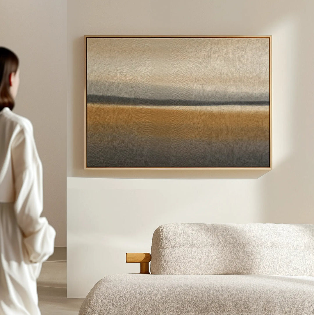 A person in a white outfit stands in a sunlit, minimal room with a beige sofa and the Honey Mesa Canvas Art, which features soft earth tones and enhances the modern home decor.