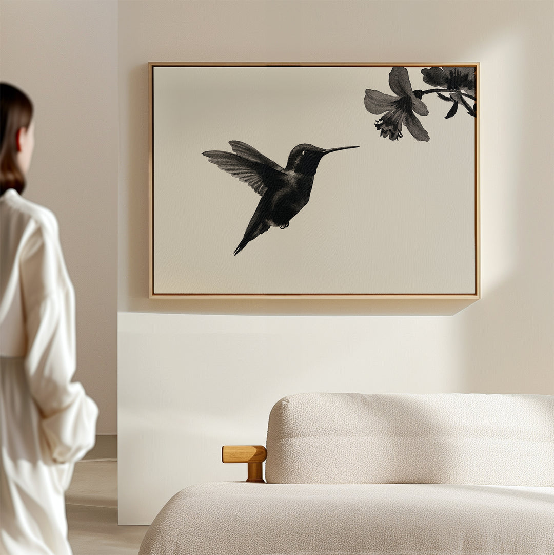 A woman stands in a light-filled, minimal room, admiring the Hummingbird Motion Canvas Art above a beige sofa—a striking minimalist piece that enhances her modern decor.