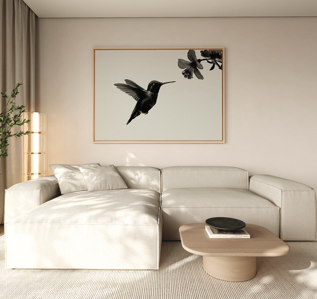 A modern living room features a light beige sectional sofa, a light wood coffee table, and the Hummingbird Motion Canvas Art—a large minimalist piece with a black hummingbird print—bathed in soft natural light that enhances the contemporary decor.