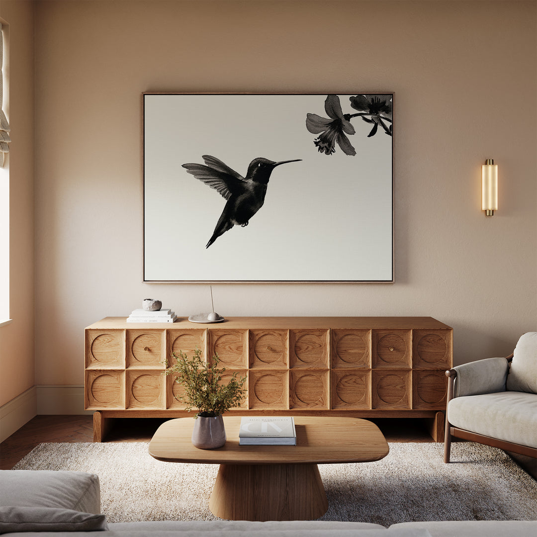 A modern living room in neutral tones features minimalist decor, including the Hummingbird Motion Canvas Art above a wooden sideboard, complemented by a sleek coffee table, vase, and light-toned sofa and armchair.