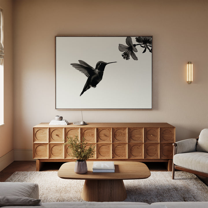 A modern living room in neutral tones features minimalist decor, including the Hummingbird Motion Canvas Art above a wooden sideboard, complemented by a sleek coffee table, vase, and light-toned sofa and armchair.