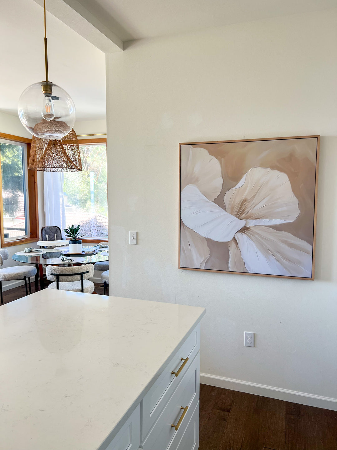 A modern kitchen with white countertops and gold-handled drawers, a glass pendant lamp, and a large abstract flower painting on the wall. A dining area with windows and natural light is visible in the background.