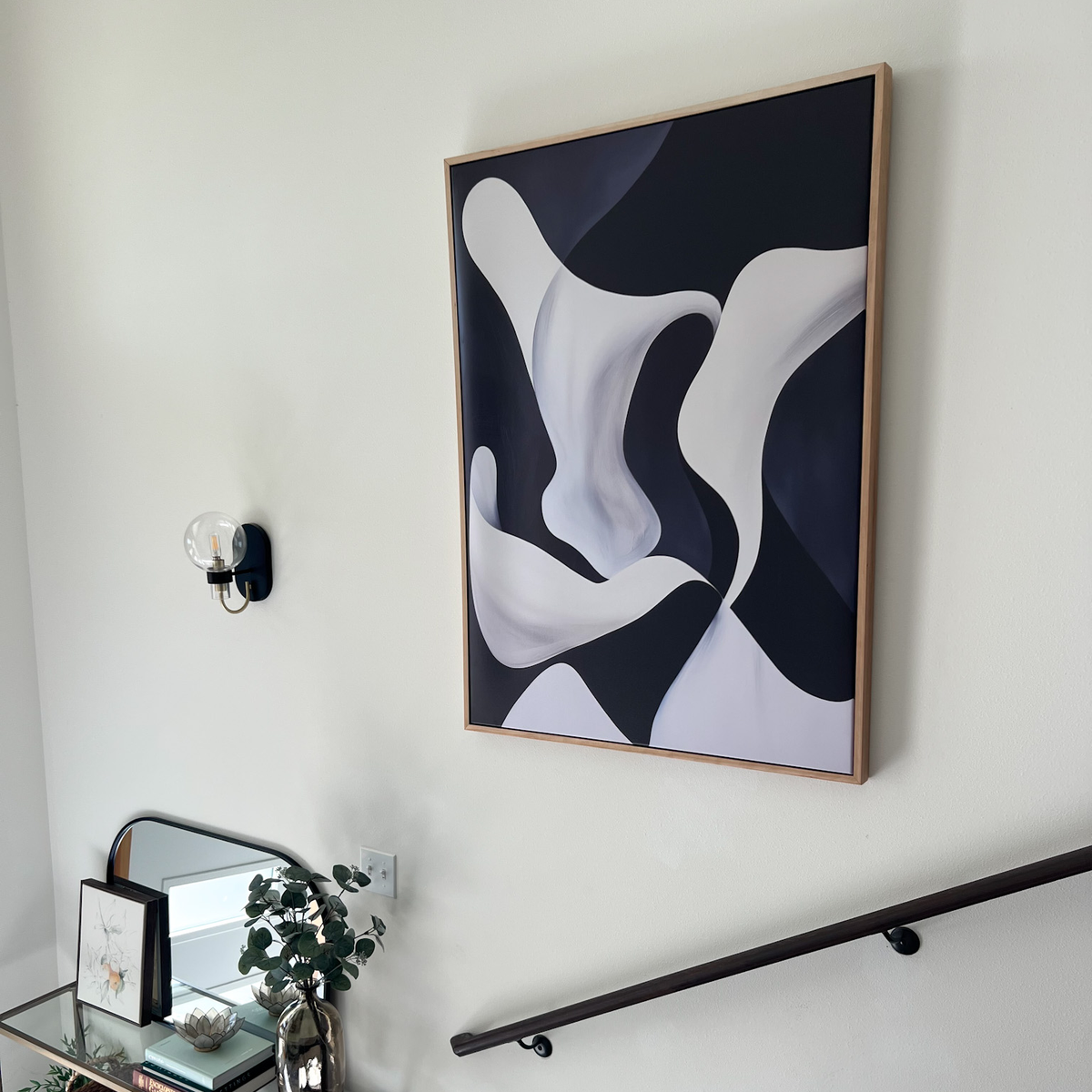 The Deep Blue Calm Abstract Canvas Art hangs above the staircase, elevating the modern space. Below, a small table with greenery, a mirror, and framed art complements the decor while a wall sconce adds stylish lighting to the living room.