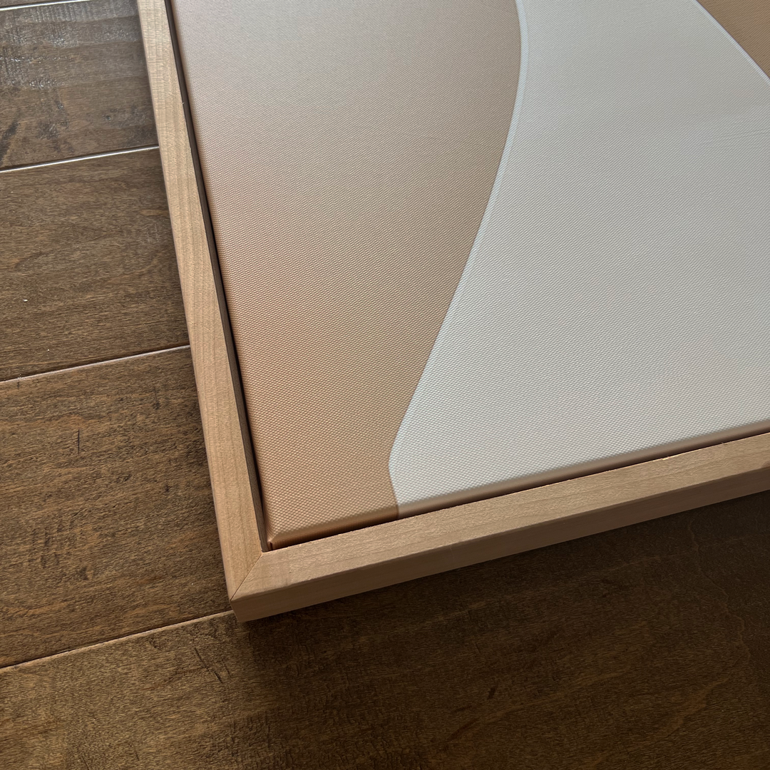 A close-up of the corner of a wooden-framed canvas featuring abstract art in beige and white tones, resting on a brown wooden floor.