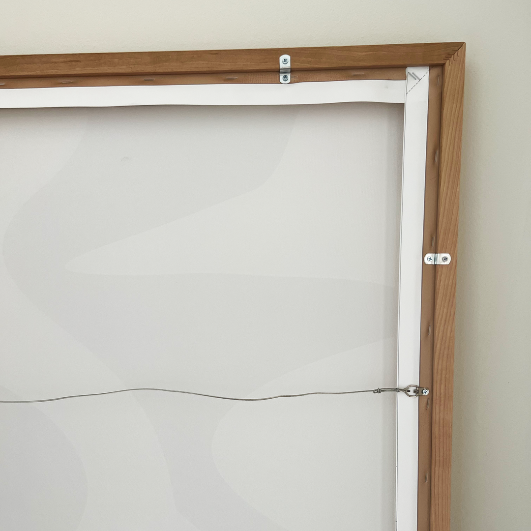 The back of a wooden picture frame with wire for hanging, attached by two screws on each side. The frame rests against a light-colored wall, with the canvas back visible.