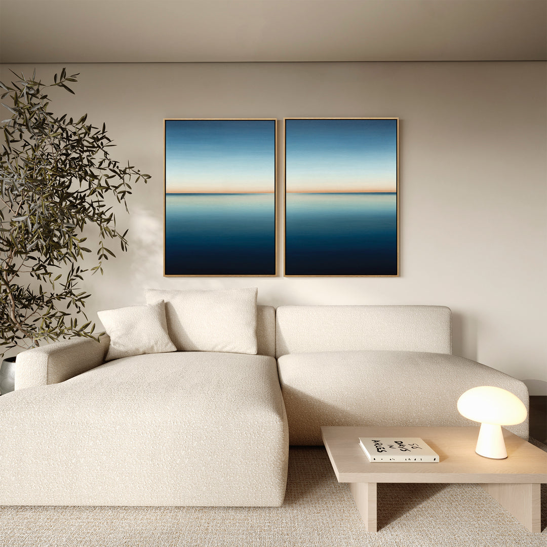 A minimalist living room with a cream sofa, a small wooden table with a lamp and book, a leafy plant, and two pieces of modern decor featuring the Indigo Passage Set of 2 abstract blue and beige wall art.