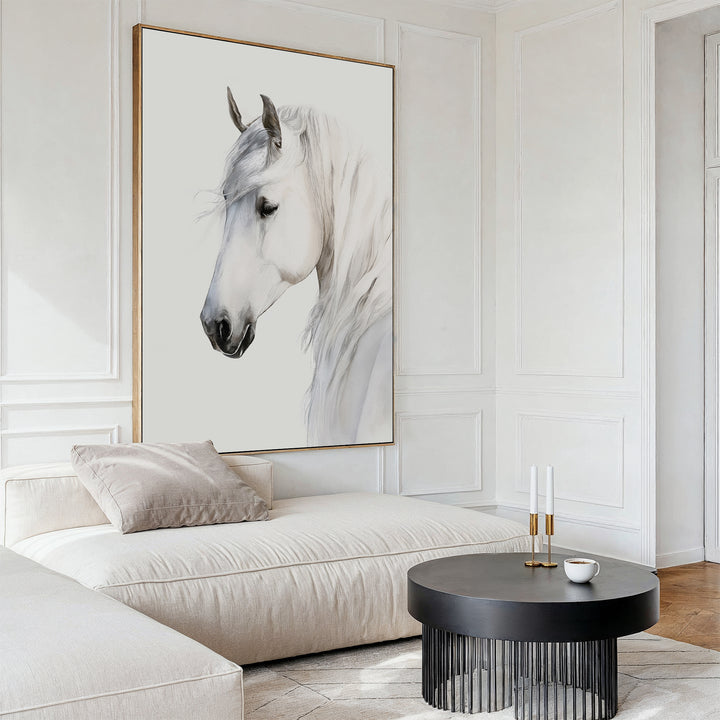 A modern living room with a large white sofa, a round black coffee table, neutral tones, minimal decor, and the Ivory Mane Canvas Art—a framed painting of a white horse with a soft background—on the wall.