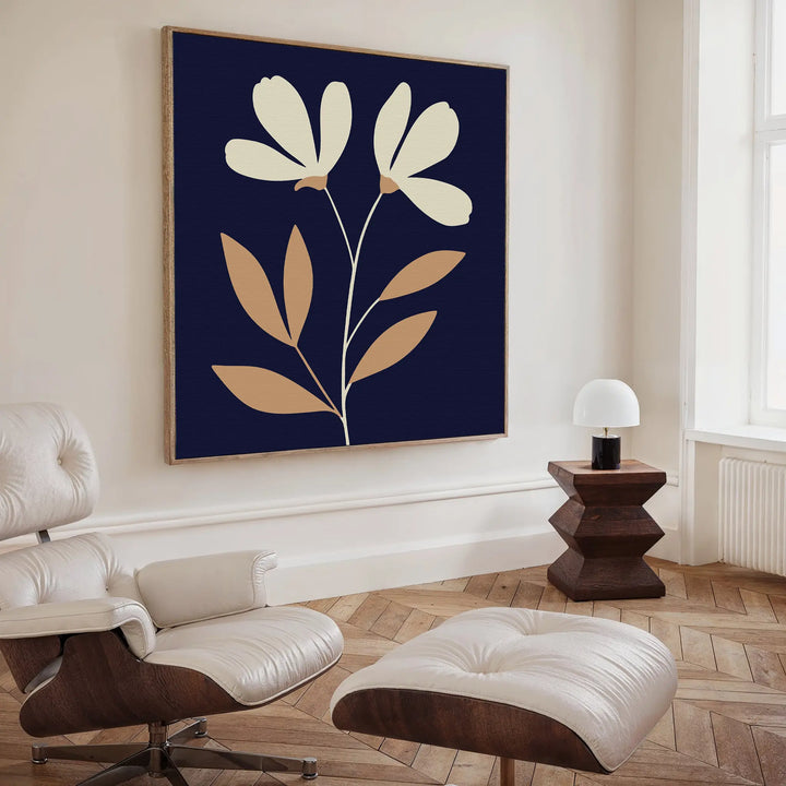 A stylish living room featuring a white lounge chair and ottoman, a wooden side table with a white lamp, and the Ivory Stems Canvas Art—large wall art displaying beige flowers and ivory stems on a dark blue background.
