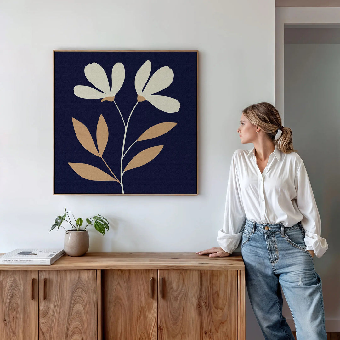 A woman in a white shirt and jeans stands by a wooden sideboard, admiring the Ivory Stems Canvas Art, which features minimalist ivory stems and beige flowers on a dark background. A small plant and books enhance the modern, white-walled room.