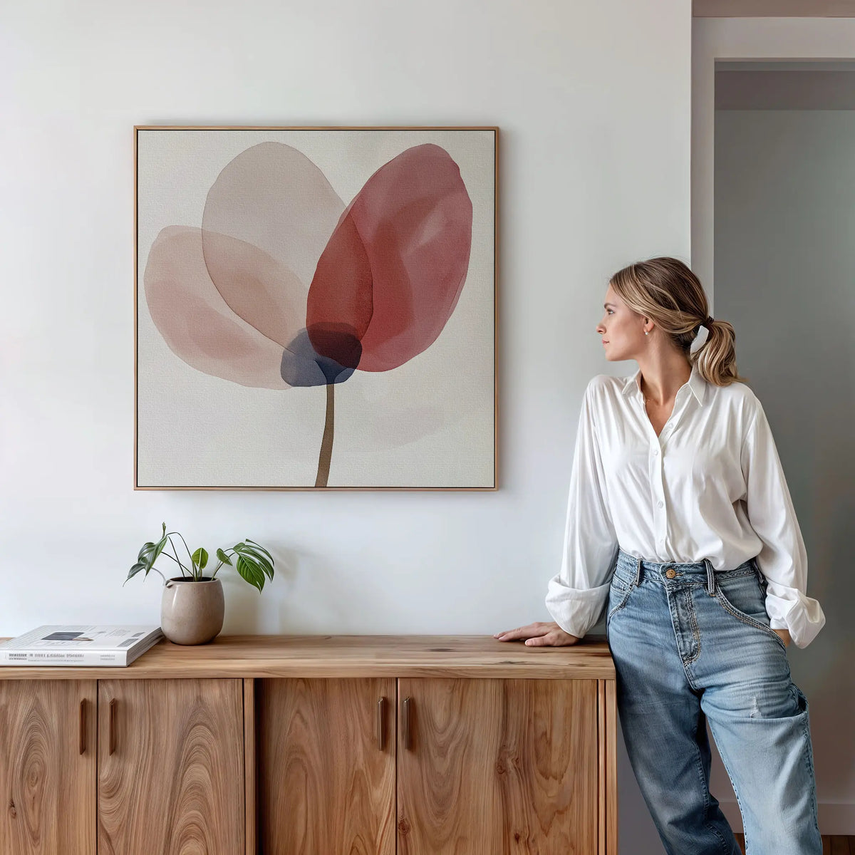 A woman in a white blouse and jeans leans on a wooden cabinet, admiring the Layered Bloom Canvas Art. A small potted plant and book rest atop the cabinet, enhancing the modern wall decor of this serene space.