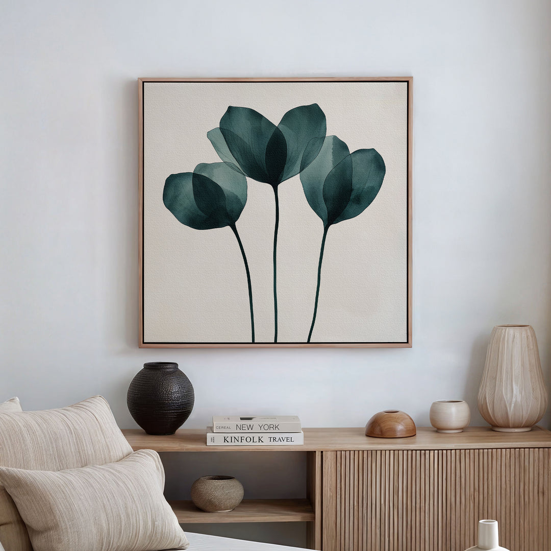 A minimalist living room with light-toned furniture features the Leafy Balance Canvas Art—a set of three abstract green leaves—framed above a wooden sideboard decorated with vases, a book, and stylish decor accents.