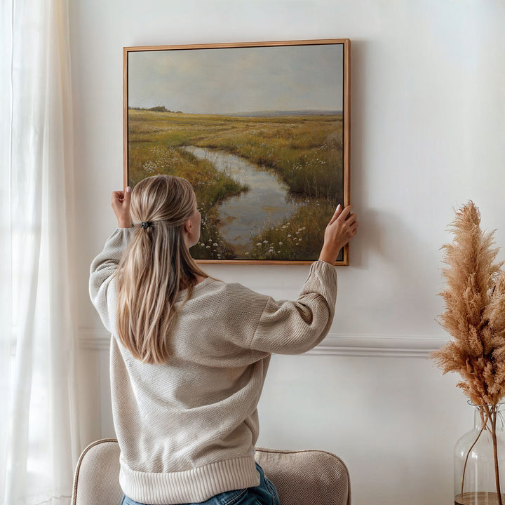 A woman with long blonde hair hangs the Marshland Stream Canvas Art—featuring a grassy field and river—on a white wall. Sunlight filters through curtains, while pampas grass in a vase adds to the stylish living room decor.