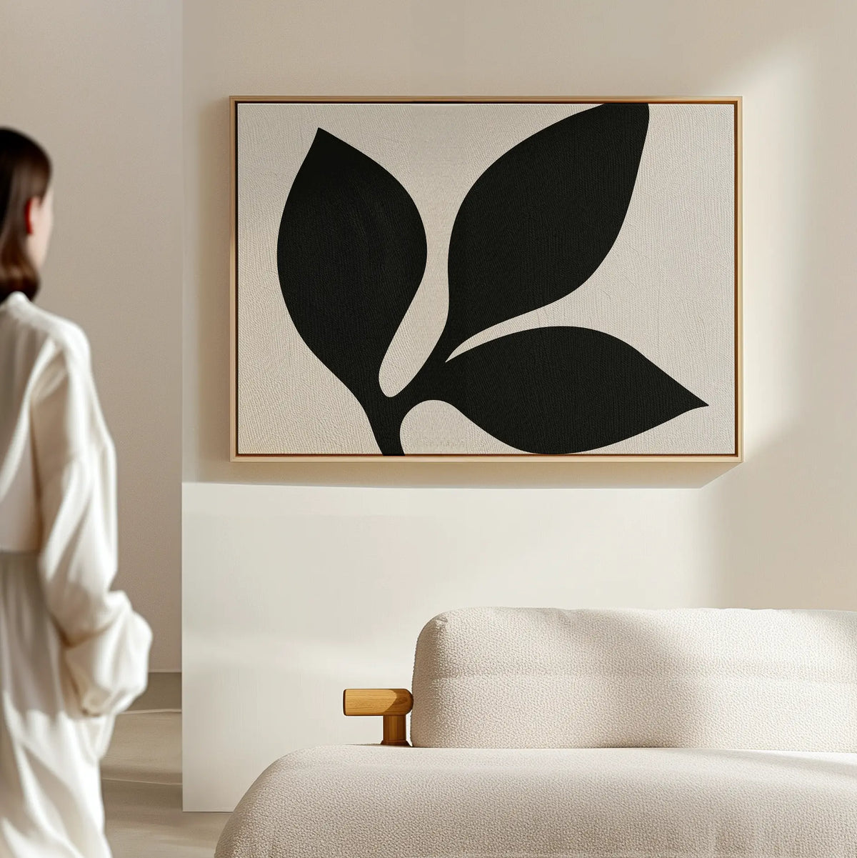 Dressed in white, a person admires the Midnight Botanist Canvas Art—featuring three striking black leaves on a light backdrop—displayed above a beige sofa, adding modern minimalist style to the living room.