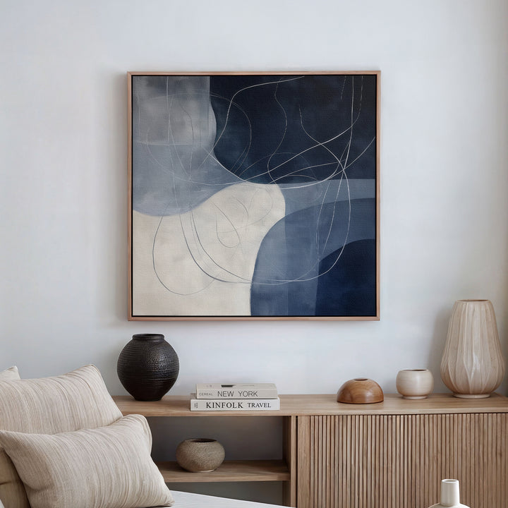 A modern living room features a beige sofa, wooden sideboard with vases and books, and the Midnight Lines Abstract Canvas Art—a large painting in dark blue, gray, and cream with white curved lines—elevating the decor.