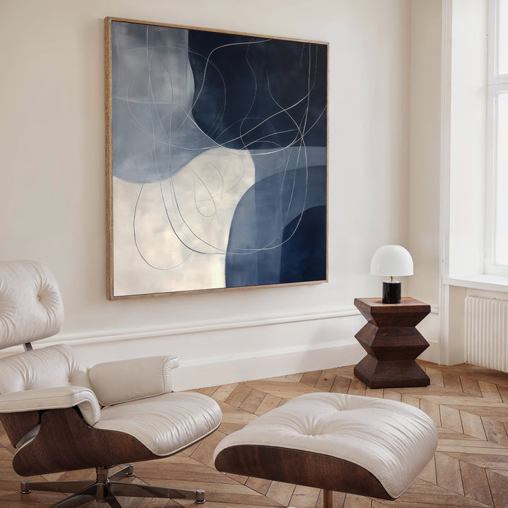 A modern living room with a cream lounge chair and ottoman, geometric side table with a white lamp, herringbone wood floors, and Midnight Lines Abstract Canvas Art—a large blue and white painting—as the striking focal point.