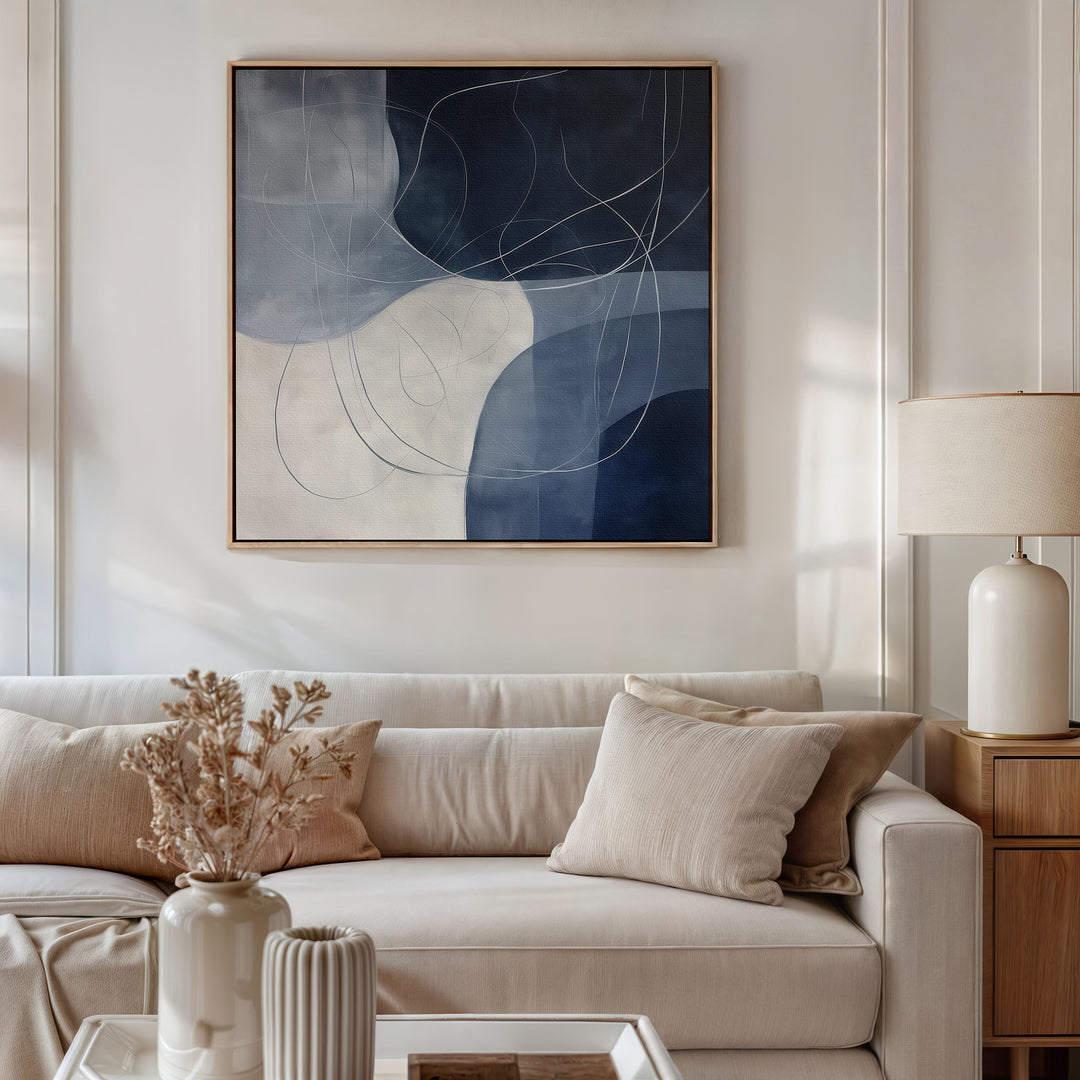 A modern living room with a beige sofa, matching cushions, a light wood side table with a ceramic lamp, dried flowers in a vase, and the elegant Midnight Lines Abstract Canvas Art completes the decor.