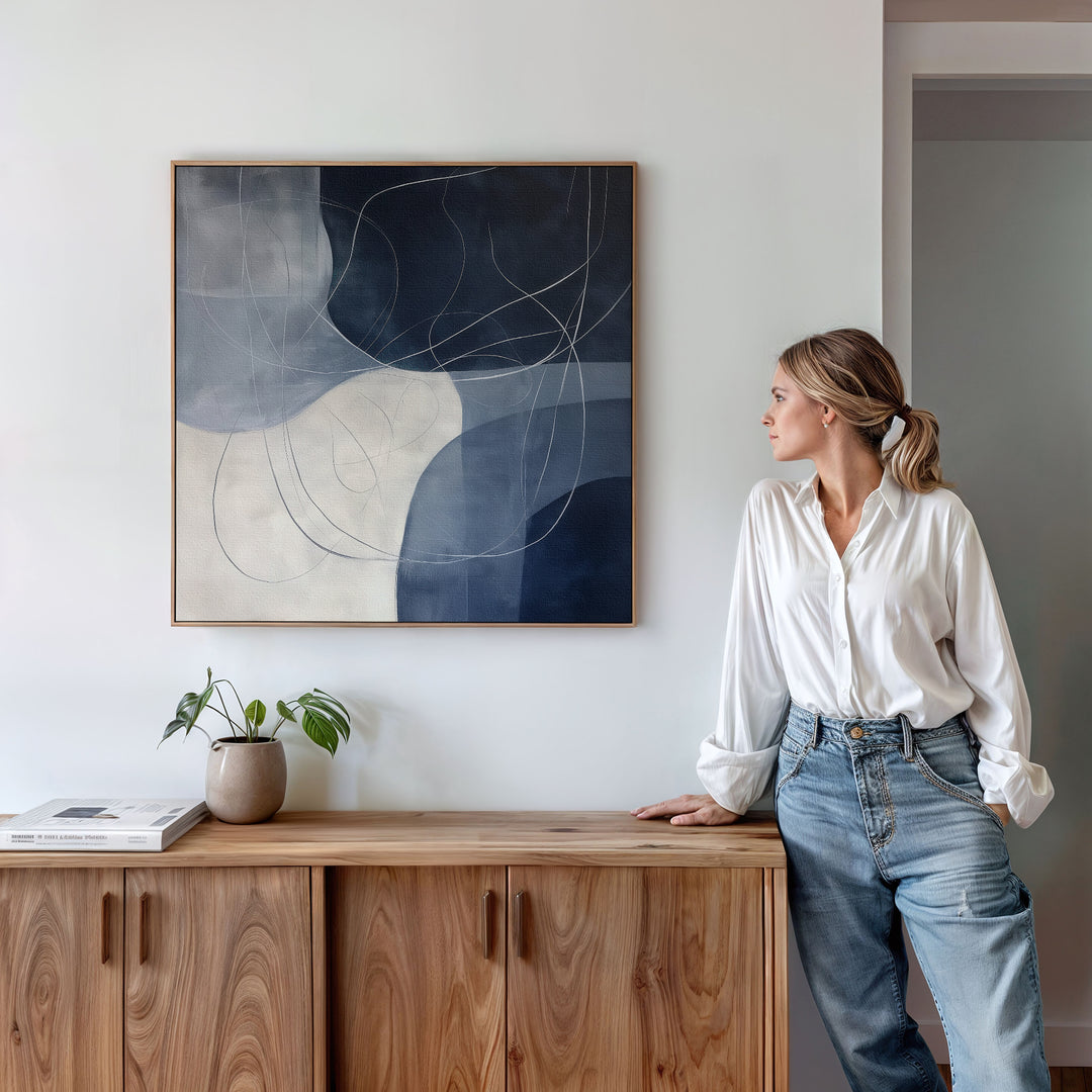 A woman in a white blouse and jeans leans on a wooden cabinet, admiring the Midnight Lines Abstract Canvas Art in blue and white. A potted plant and books complete the modern, minimalist vibe of the living room.