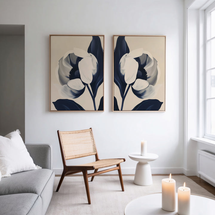A modern living room features a wooden chair, round side table, gray sofa, and three lit candles. Above the chair hang the Midnight Tulip Set of 2 botanical wall art in navy and cream, adding style as natural light fills the space.