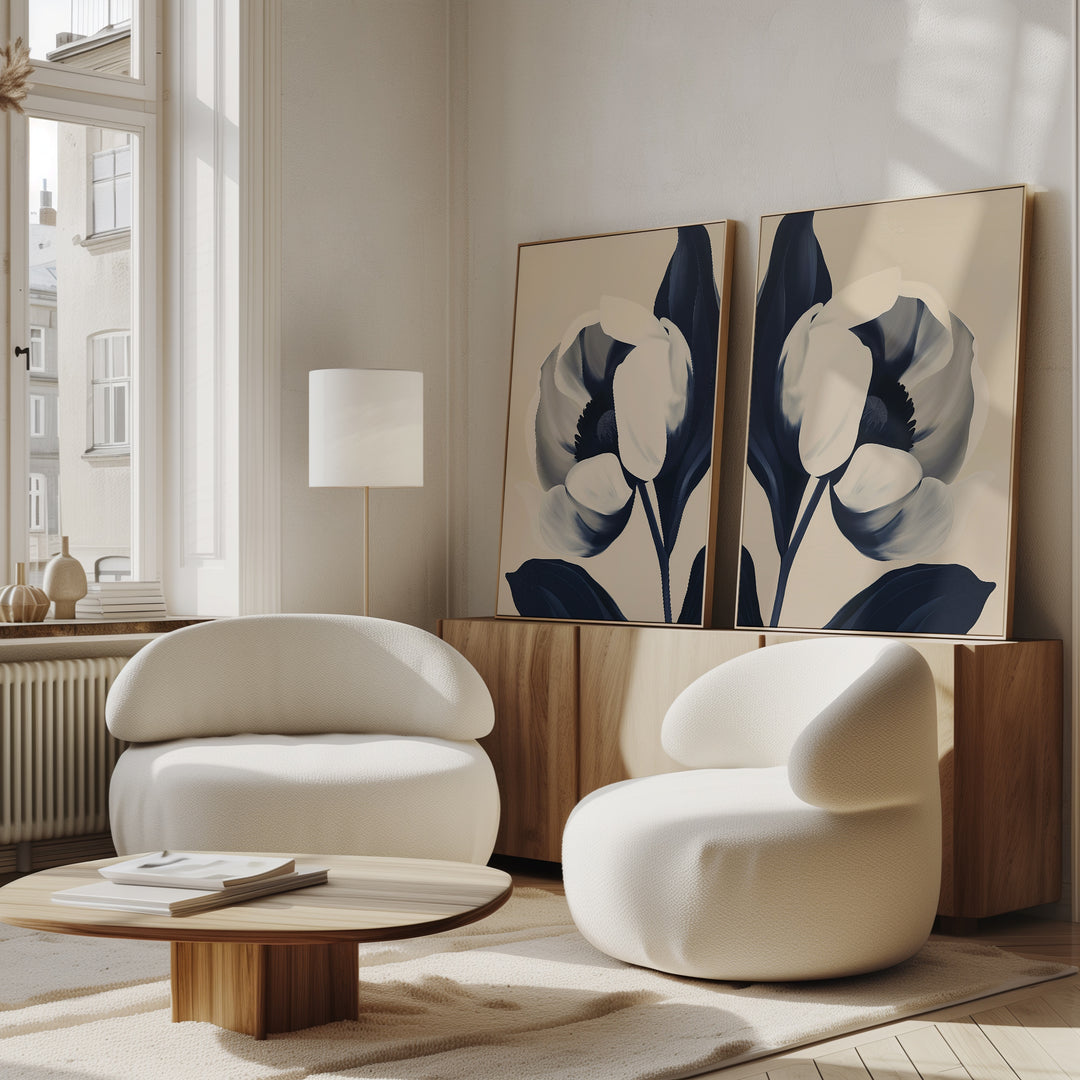 A modern living room features two white rounded chairs, a low wooden coffee table, and a sideboard displaying the Midnight Tulip Set of 2 minimalist floral canvas artworks. Sunlight from tall windows creates a cozy, inviting atmosphere.