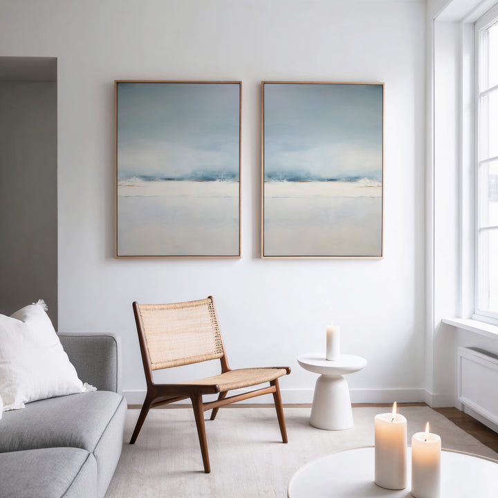 A minimal, modern living room with a wooden chair, a white side table holding a lit candle, and a grey sofa with white cushions. Above the sofa, the Misty Morning Set of 2 adds tranquility with minimalist abstract landscape art.