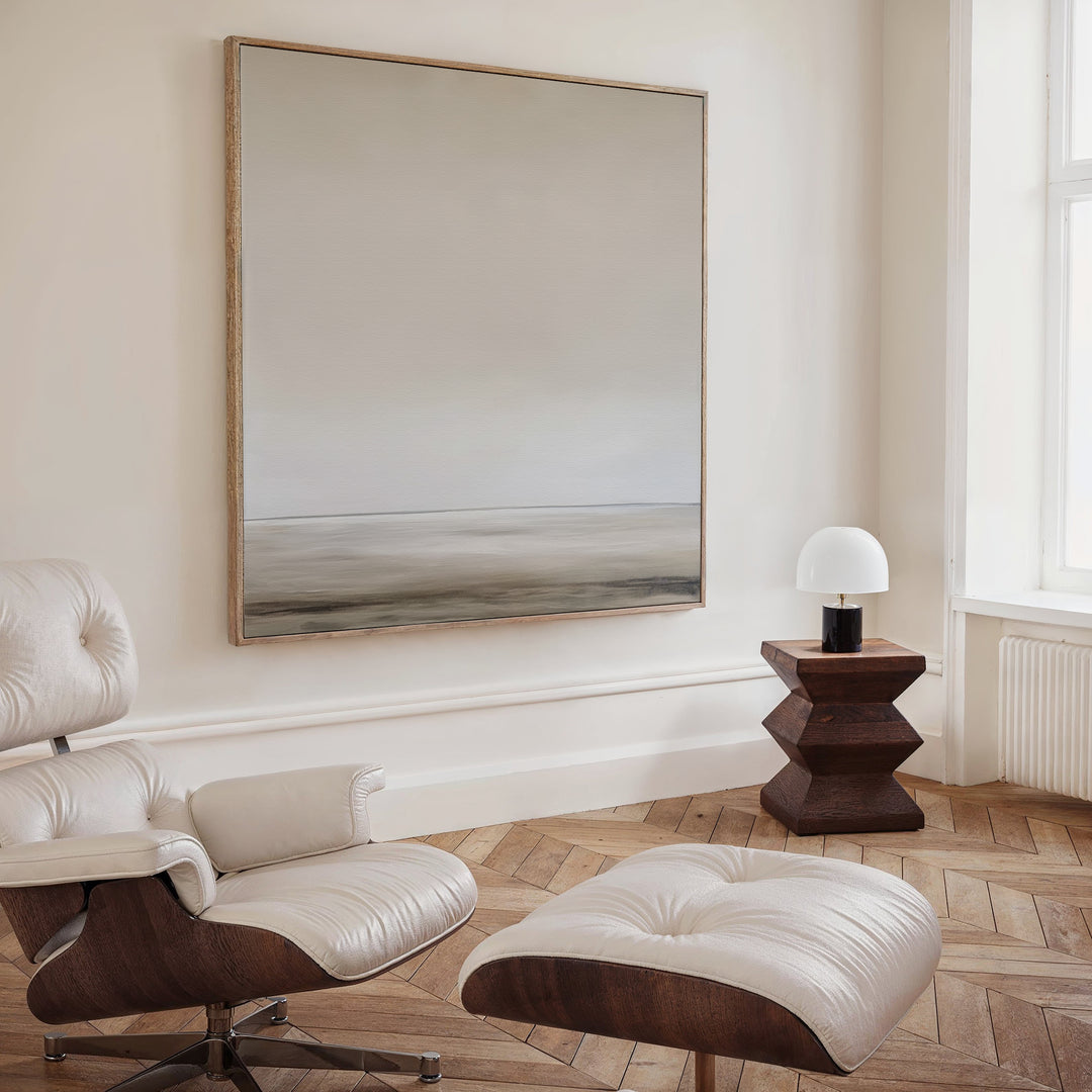A modern, minimalist room features a cream Eames lounge chair with ottoman, a geometric wooden side table with dome-shaped lamp, herringbone wood floors, and the Morning Mist Abstract Canvas Art—a neutral-toned piece—on the wall.