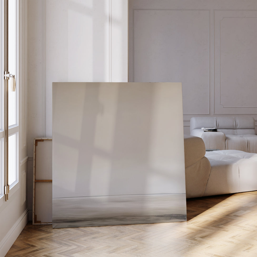 The Morning Mist Abstract Canvas Art, featuring a soft neutral seascape, leans against the wall in a sunlit modern living room with parquet floors and white furniture.