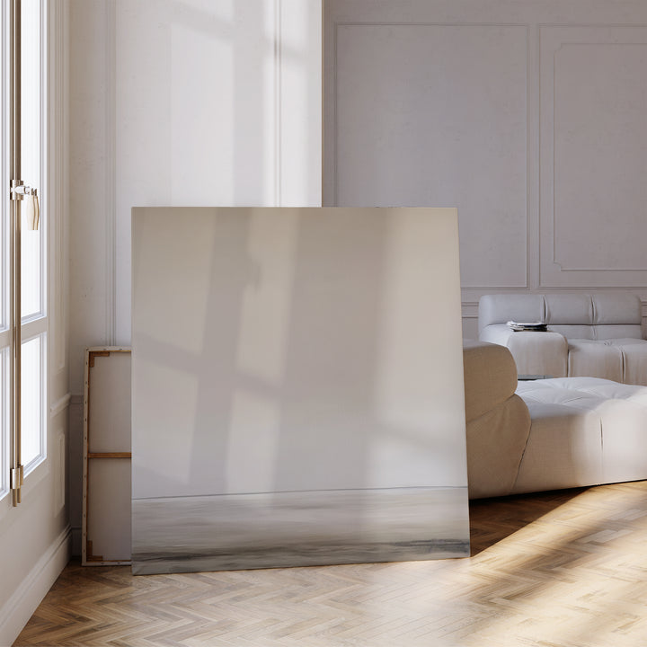 The Morning Mist Abstract Canvas Art, featuring a soft neutral seascape, leans against the wall in a sunlit modern living room with parquet floors and white furniture.