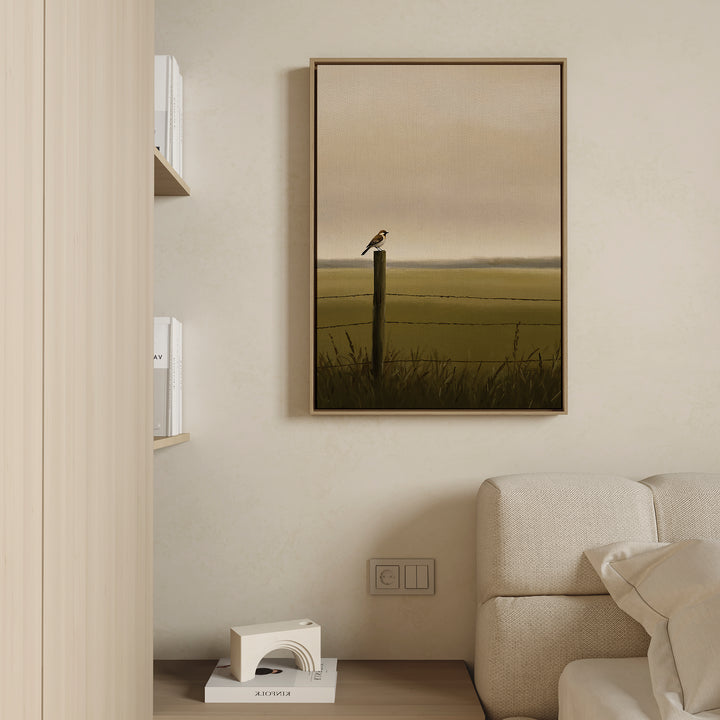 A minimalist living room corner with a beige sofa, shelves of books, and the Morning Perch Canvas Art—a framed painting of a bird on a fence post—adds understated charm to earthy interiors.