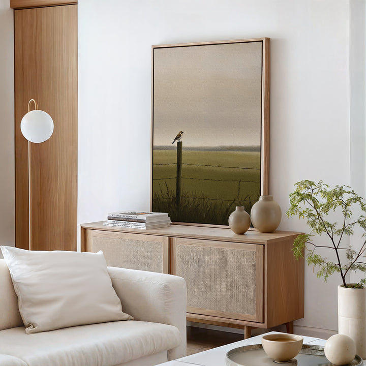 A minimalist living room features a light wood sideboard, neutral decor, potted plant, and the Morning Perch Canvas Art—a framed bird painting. Earthy elements blend with a modern floor lamp and white sofa for a serene vibe.