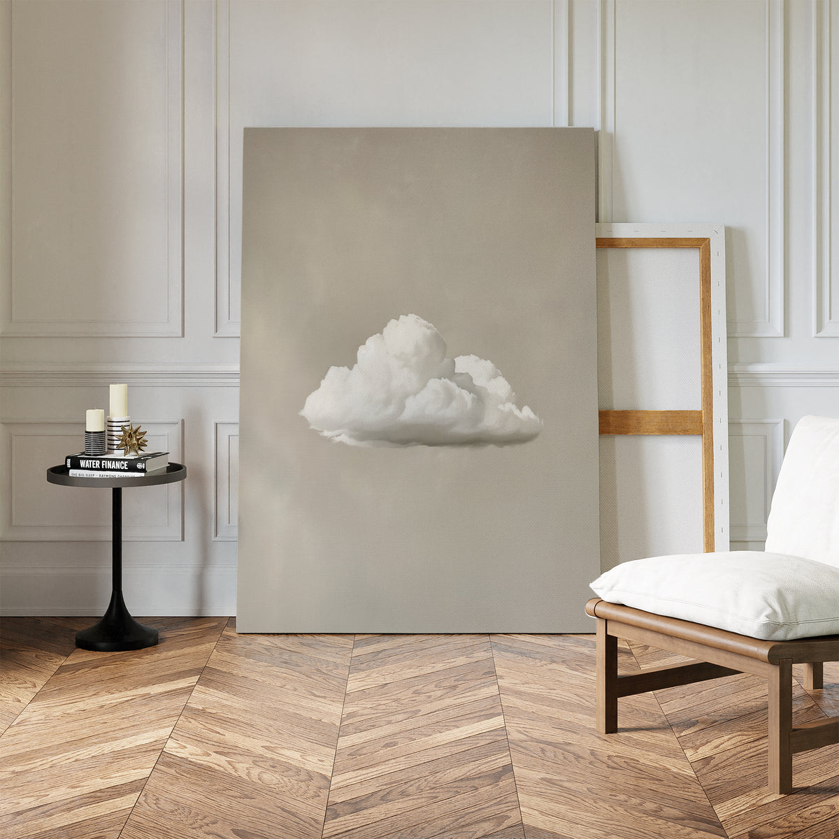 The Morning Sky Abstract Canvas Art, featuring a white cloud on beige, leans beside an empty frame. A white cushioned chair and black side table with candles complete the serene decor on the herringbone wood floor.