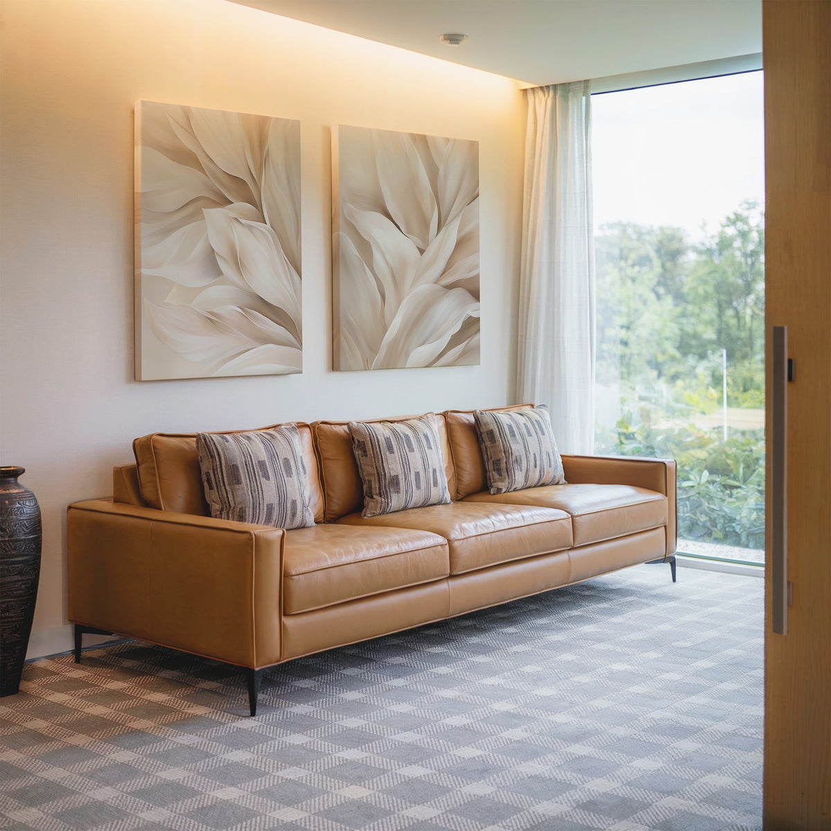 A modern living room with a tan leather sofa, four patterned cushions, the Natural Texture Set of 2 abstract leaf paintings as wall decor, a large window with sheer curtains, and a dark vase on the side.