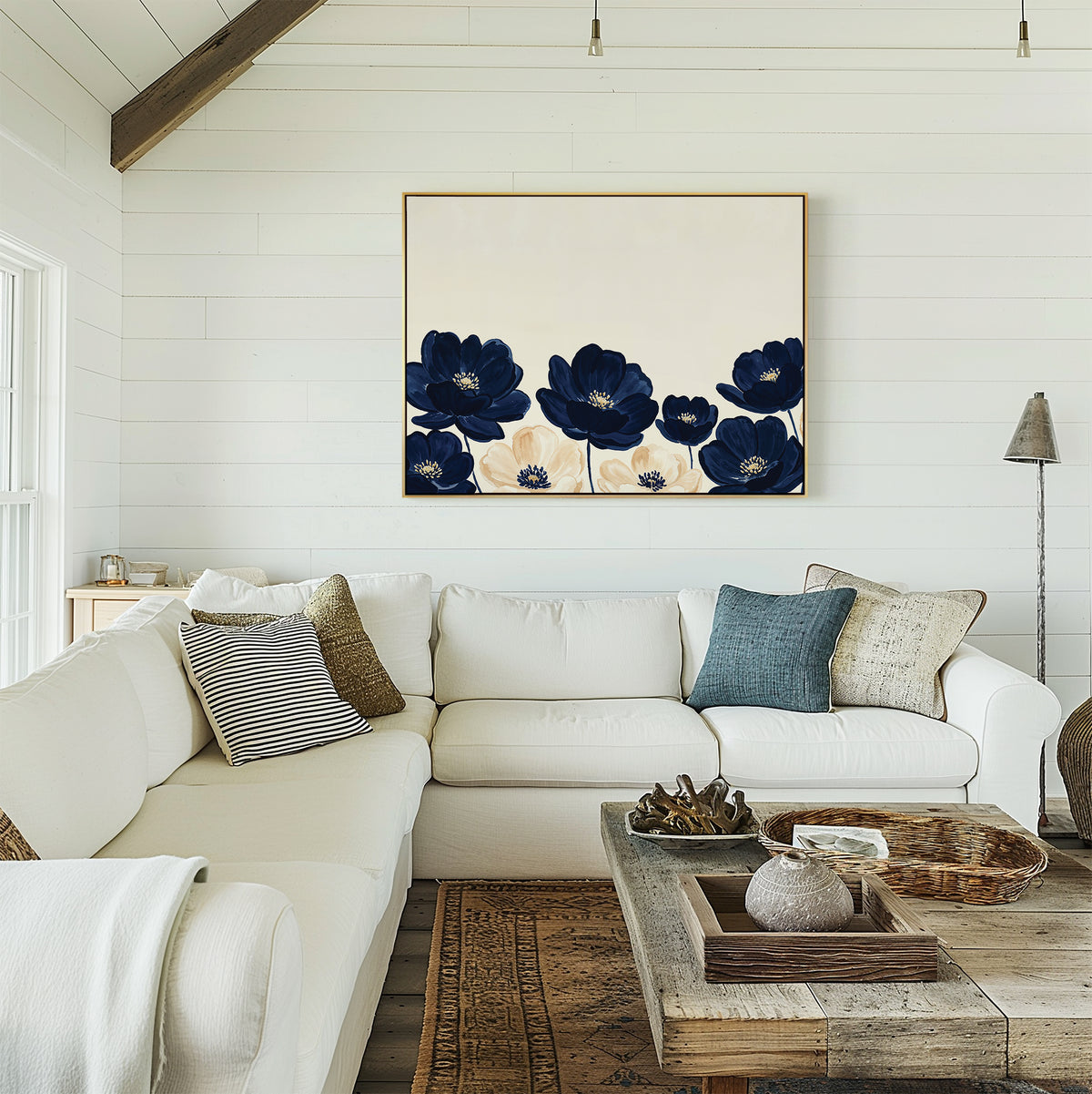 A cozy living room features a white sectional sofa, assorted pillows, a wooden coffee table, and the Nightshade Canvas Art – Horizontal with blue and white floral accents. White shiplap walls and rustic decor complete the inviting space.