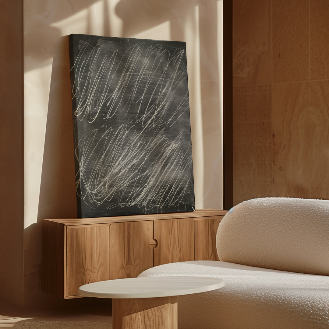 The Echoes in Chalk Abstract Canvas Art, featuring minimalist white markings on black, leans against a beige wall atop a wooden cabinet beside a textured white sofa and round coffee table in a sunlit room.