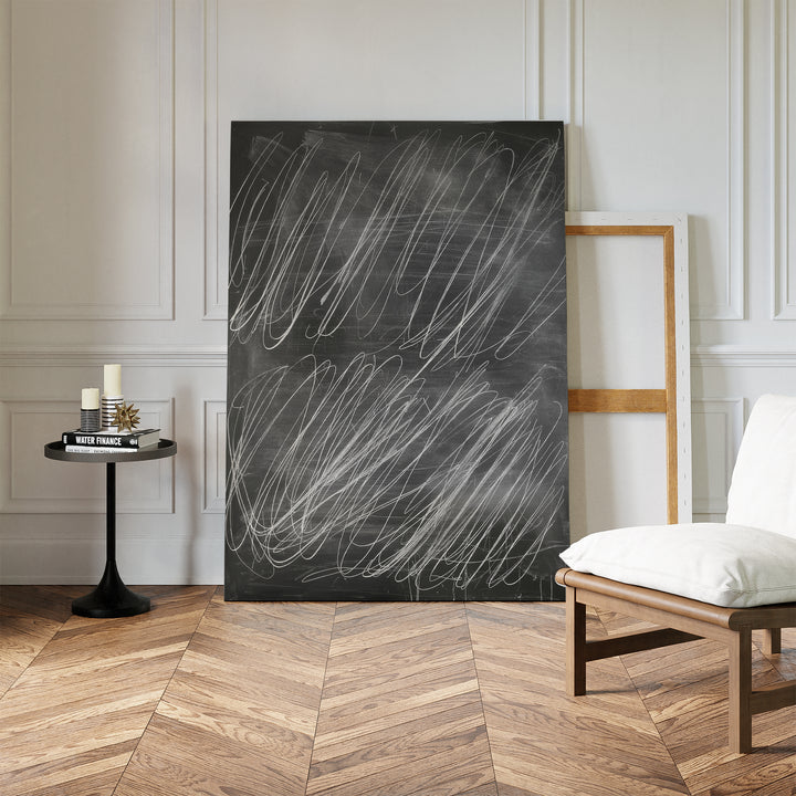 The Echoes in Chalk Abstract Canvas Art leans against a white paneled wall beside an empty frame, while a cushioned chair and a small table with candles and books sit on the herringbone floor.