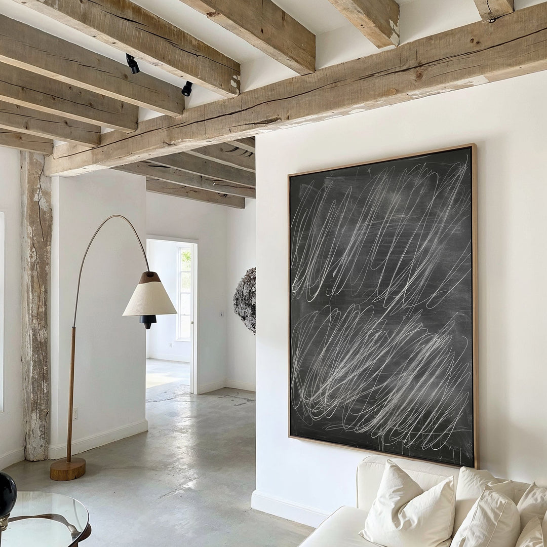 A modern living room with exposed wooden beams, white walls, a white couch with pillows, a curved floor lamp, and Echoes in Chalk Abstract Canvas Art featuring white scribbles as the focal wall decor.