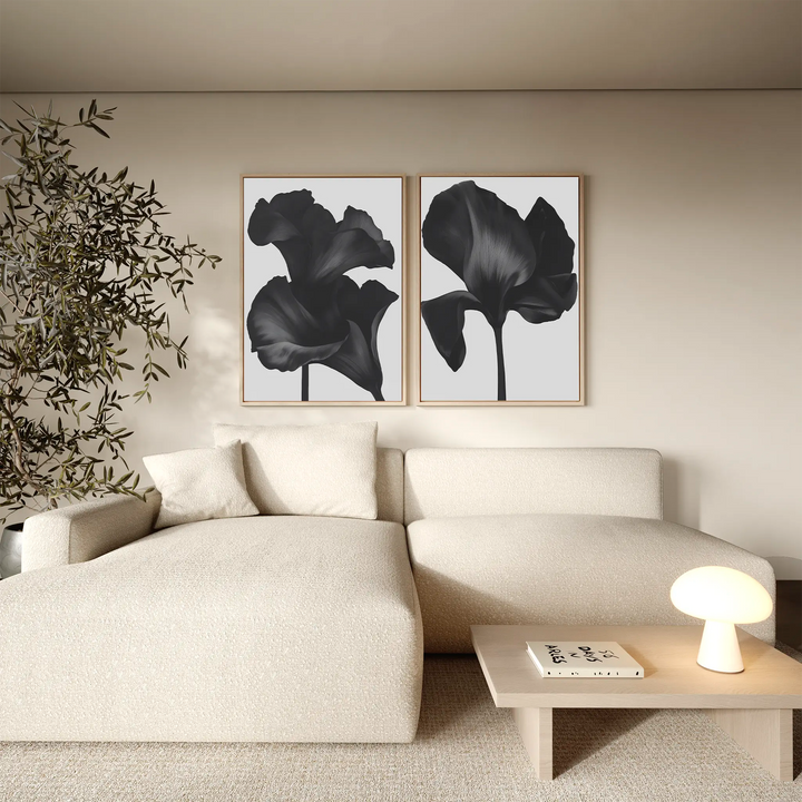 A minimalist living room with a beige sofa, light wooden table, small lamp, potted plant, and the Noir Bloom Set of 2 wall art prints hanging above the sofa.