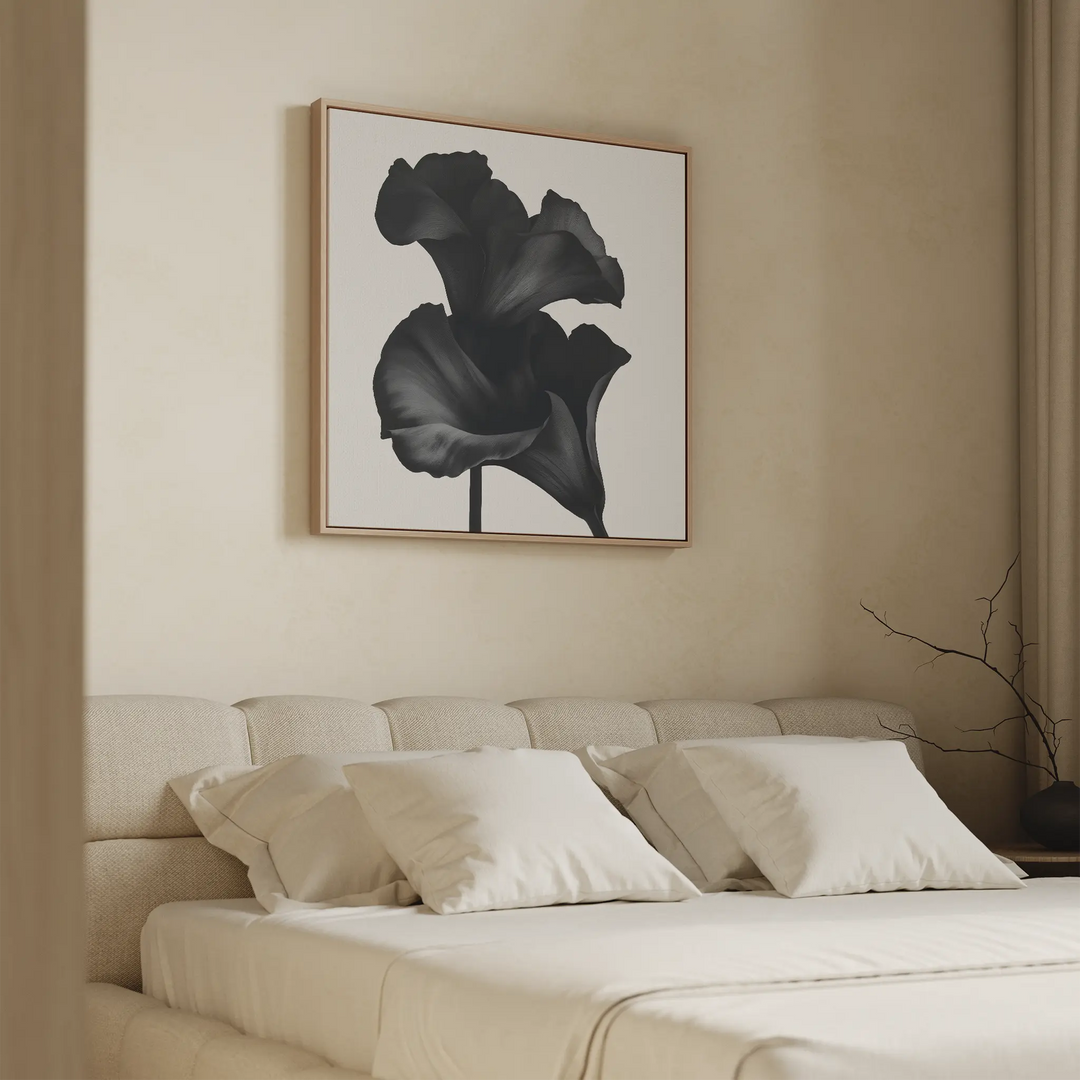 A modern bedroom features a beige upholstered bed with cream bedding and four pillows. Above the bed hangs the Noir Bloom Canvas Art - Square, showcasing a black flower. A minimal branch in a vase rests on the nightstand.